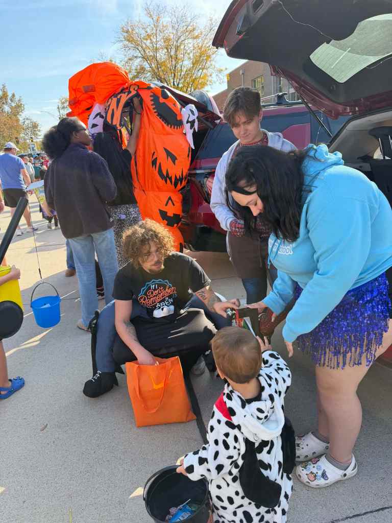 A festive scene at an outdoor event, where children and adults are engaging in activities. A toddler dressed in a Dalmatian costume is interacting with a woman in a blue hoodie while another individual sits nearby. Colorful Halloween decorations are visible, including an inflatable pumpkin. The setting is lively, with people enjoying the day.