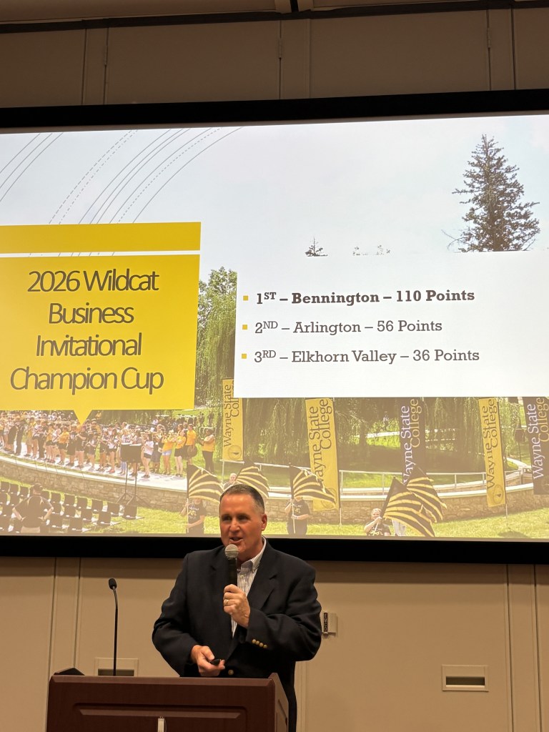 A speaker presenting at the 2026 Wildcat Business Invitational Champion Cup event, with a scoreboard showing Bennington in 1st place, Arlington in 2nd, and Elkhorn Valley in 3rd. The background features an outdoor scene with trees and banners.