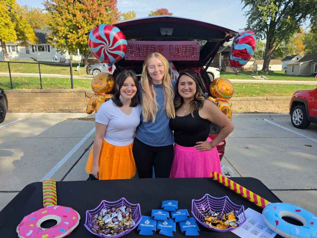 Three women posing together at a festive event, wearing colorful skirts. A table in front of them displays candy and decorative items, with a trunk decorated with balloons in the background.