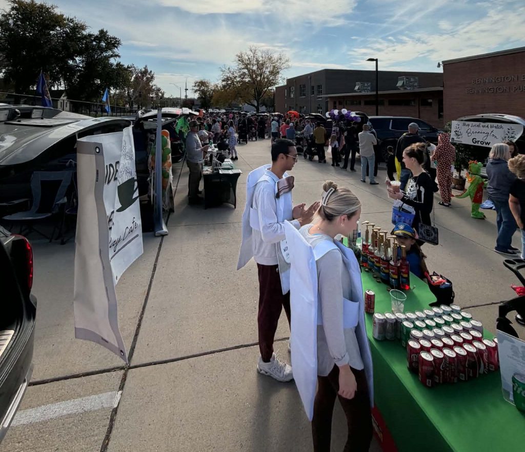 Two participants in white lab coats stand at a vendor table displaying drinks and trophies during a community event. People are gathered in the background, with some interacting and others walking by.