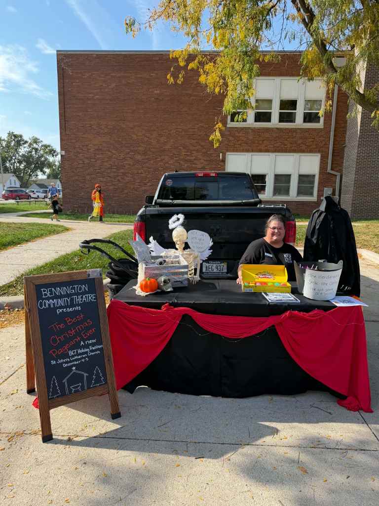 A woman sitting behind a table with a black tablecloth and red accents, promoting 'The Best Christmas Pageant Ever' for the Bennington Community Theatre, with decorations including a skeleton and pumpkin.
