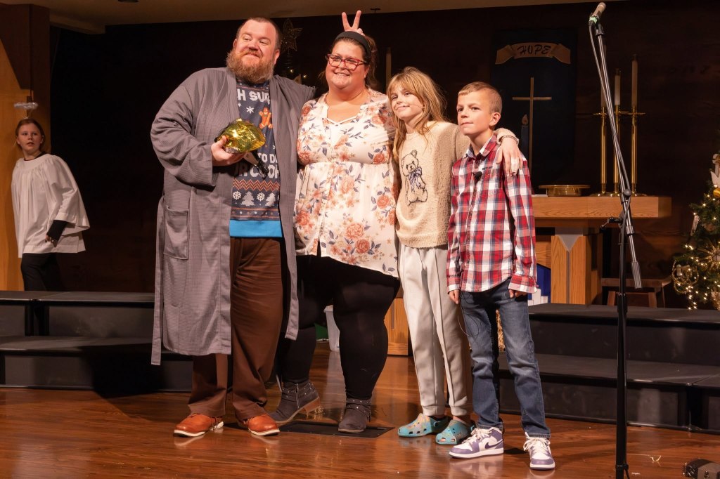 A group of four people posing on stage, smiling at the camera. One man in a robe holds a gift, while three others, including a woman and two children, stand together. A girl in the background watches.