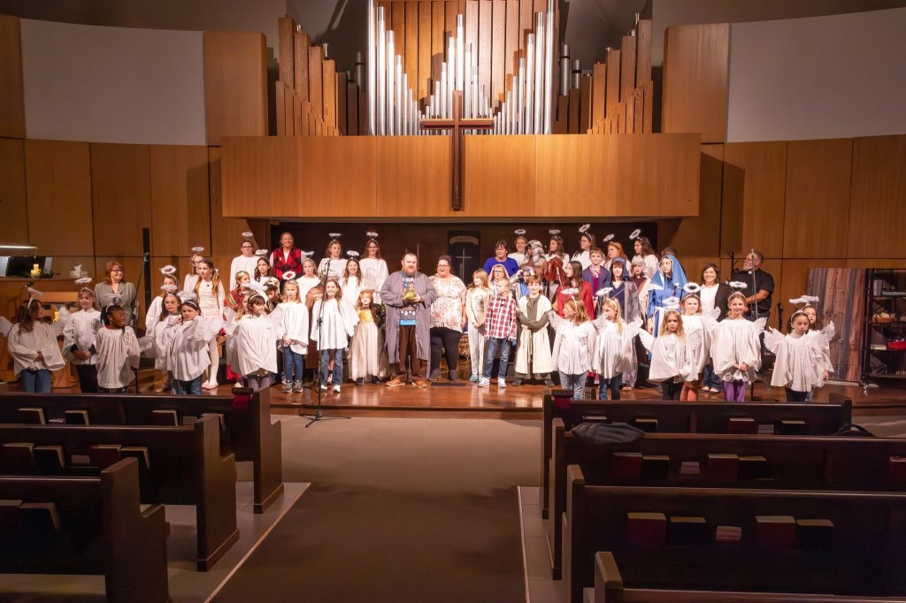 A large group of children and adults dressed in various costumes, including white robes and angel wings, gathered on a stage in a church setting. Behind them is a large pipe organ and wooden panels, with audience seating visible in the foreground.
