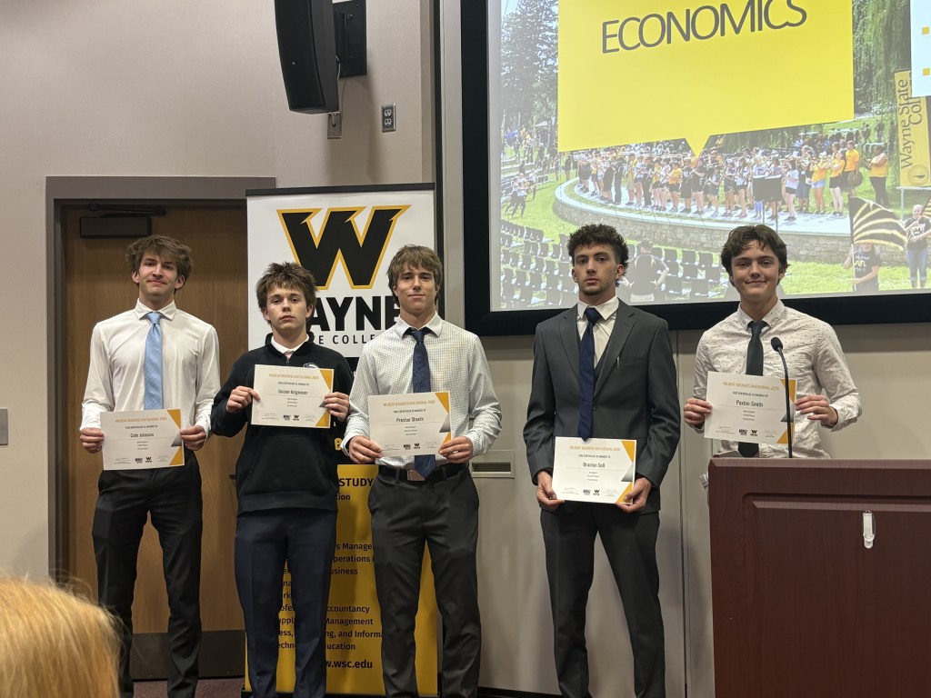 Five students standing in formal attire, holding certificates at a recognition event, with a banner in the background displaying 'Wayne State College' and the word 'Economics'.