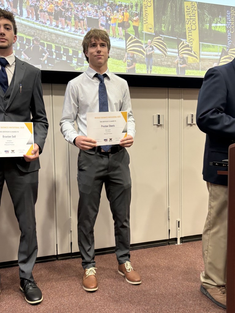Two young men stand on stage, each holding a certificate. The man on the left, Braxton Soll, is dressed in a black suit and tie, while the man on the right, Preston Sheets, wears a white shirt with a light blue tie and gray trousers. A crowd is visible in the background, along with banners for Wayne State College.