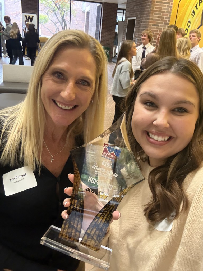Two women pose for a selfie holding an award. The woman on the left has long blonde hair, while the woman on the right has wavy brown hair. Both are smiling in a busy indoor setting with people in the background.