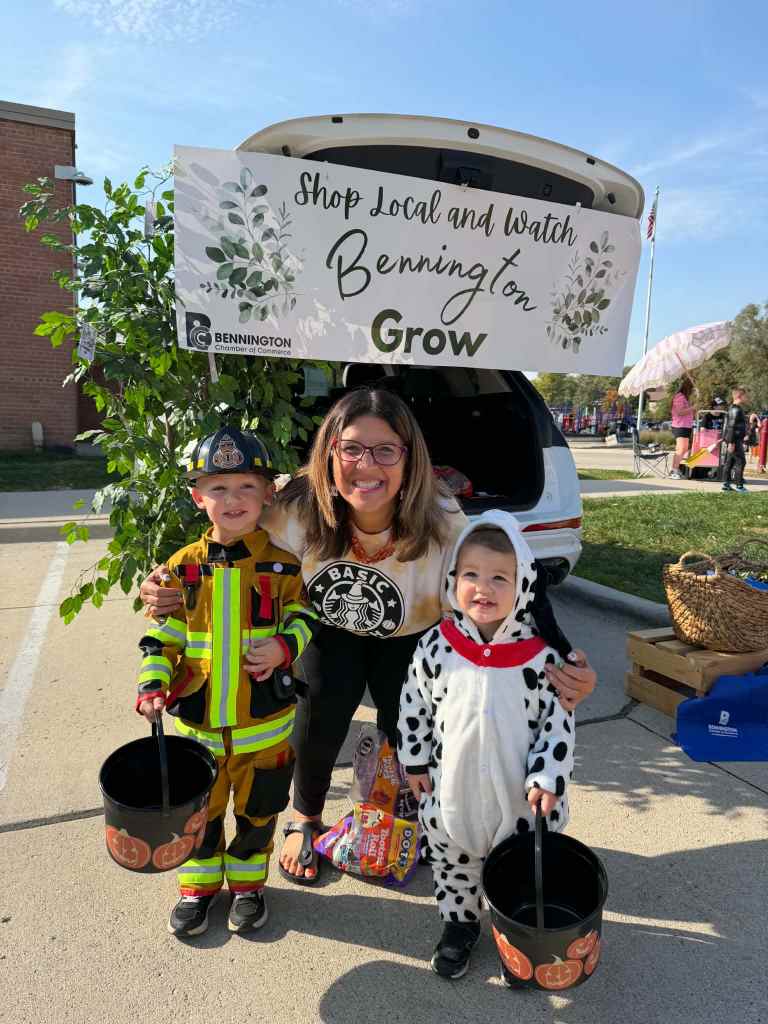 A woman with sunglasses stands between two children dressed in Halloween costumes, one as a firefighter and the other as a dalmatian. They are holding black Halloween buckets and smiling in front of a sign that says 'Shop Local and Watch Bennington Grow.'