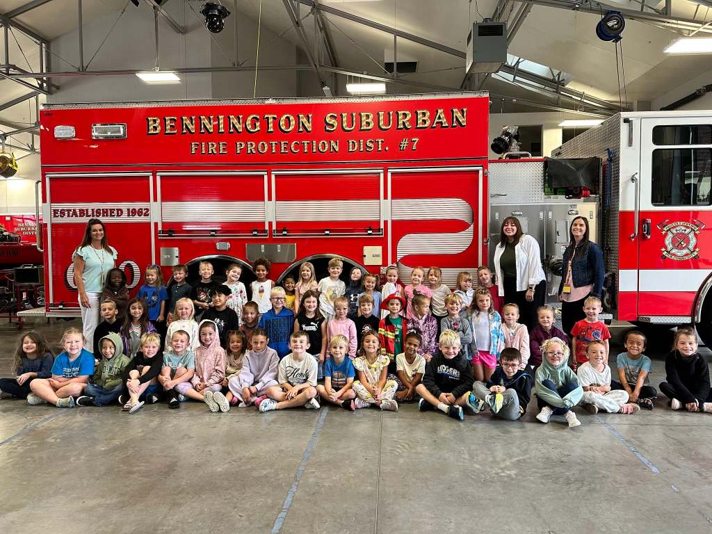 A group of children and adults posing in front of a fire truck in a fire station. The truck is red with the words 'Bennington Suburban Fire Protection Dist. #7' displayed prominently.