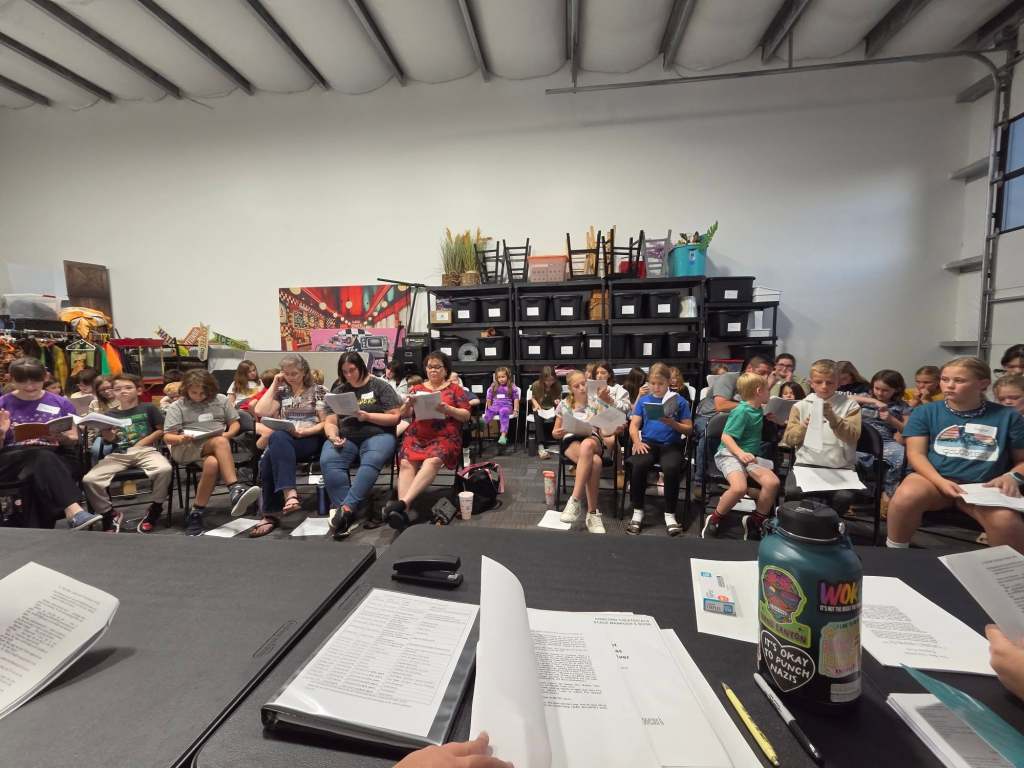 A group of children and adults seated in a rehearsal setting, reading from scripts, with various props and storage bins in the background.