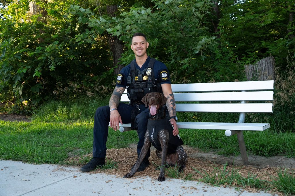 A police officer seated on a bench with a trained dog, both looking relaxed in a green park setting.
