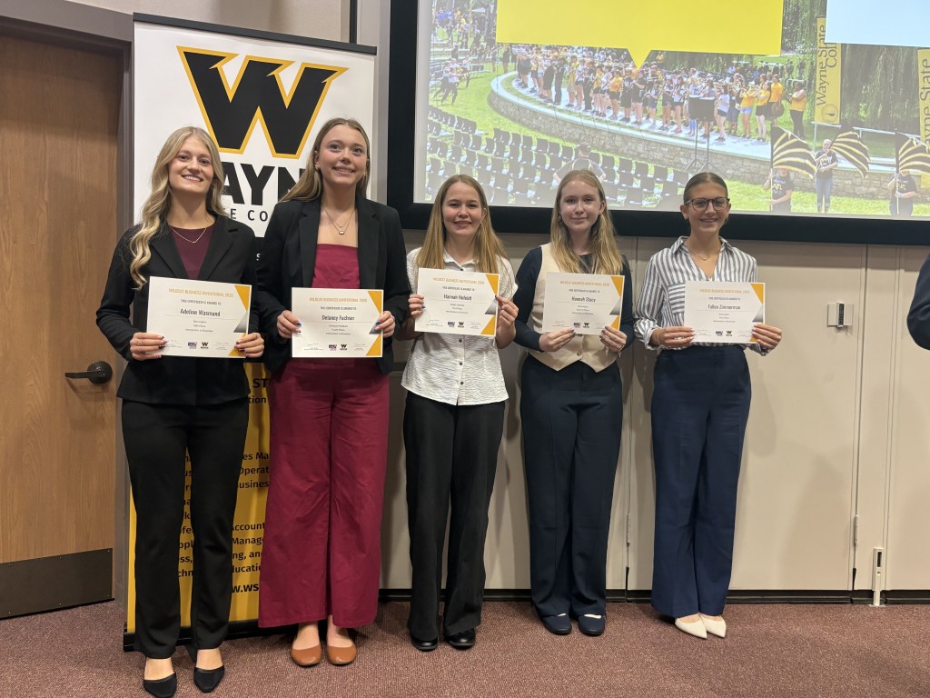 A group of five women stand together holding certificates at an award ceremony, smiling and dressed in formal attire. The background features a banner with the letter 'W' and an image of a graduation ceremony.
