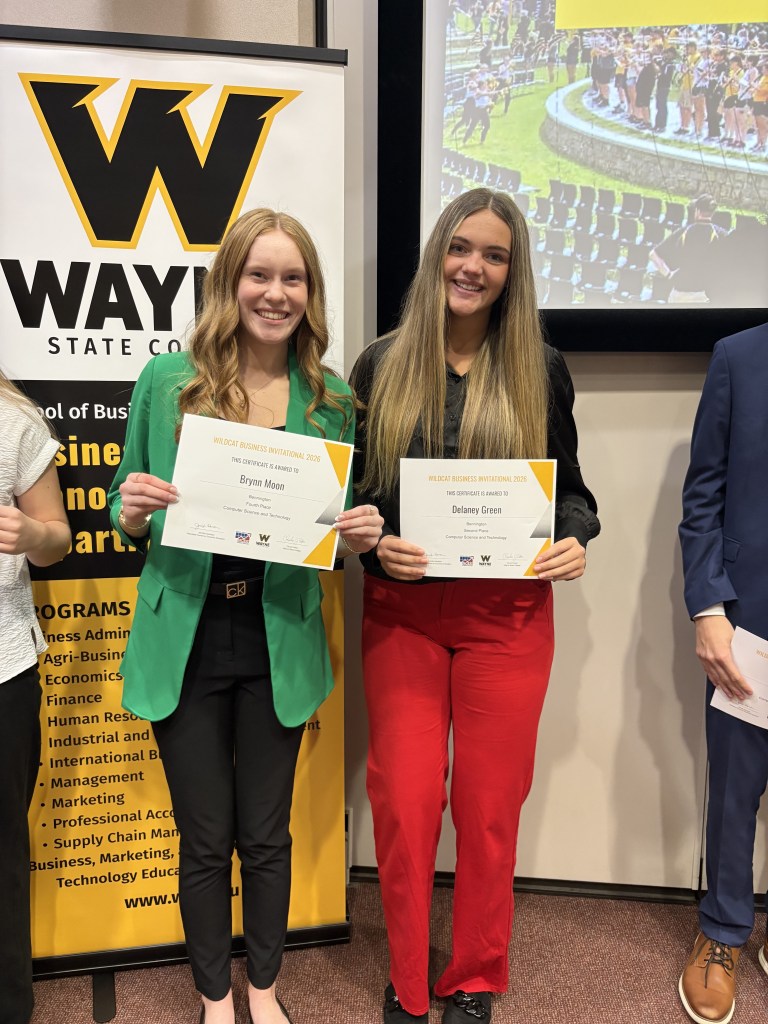 Two students proudly holding their certificates at the Wildcat Business Invitational 2023, standing in front of a Wayne State College banner.