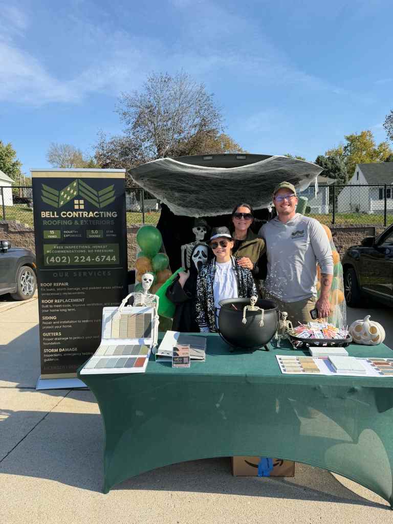 A promotional event booth for Bell Contracting featuring three people, a skeleton decoration, and various roofing materials. The booth is decorated for Halloween with a cauldron and colored balloons.