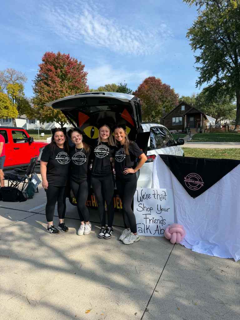 Four women in matching black shirts posing in front of a decorated car trunk at an outdoor event, with colorful trees in the background and a sign that reads 'We're that Shop Your Friends Talk ADs'.