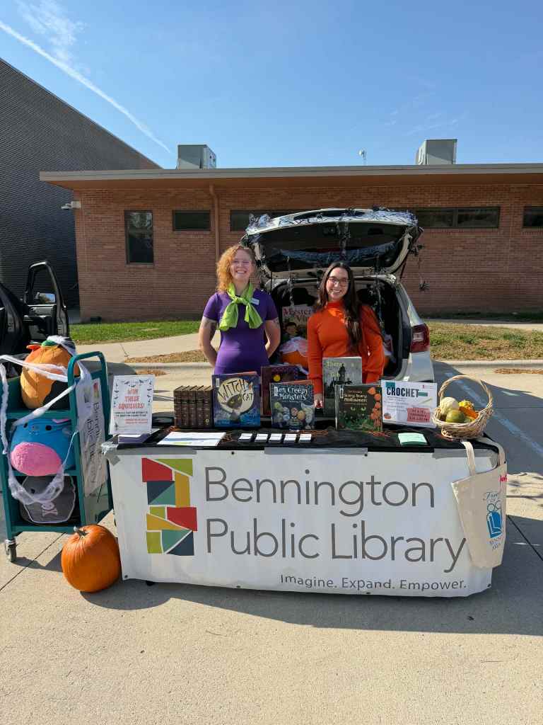 Two women are standing at a table set up for the Bennington Public Library, displaying books and crafts, with a pumpkin on the ground and a sunny sky in the background.