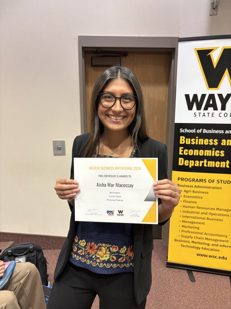 A young woman with long dark hair, wearing glasses and a black blazer, smiles while holding a certificate that reads 'Wildcat Business Invitational 2026' awarded to her for fourth place in Personal Finance.