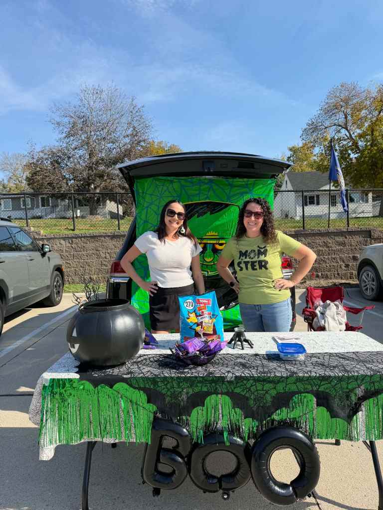 Two women standing in front of a decorated car trunk with a Halloween theme, featuring a black table with snacks, a pumpkin, and a 'BOO' balloon.