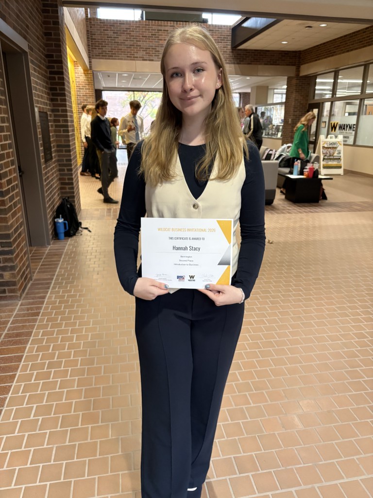 A young woman holding a certificate for the Wildcat Business Invitational 2026, smiling while standing in a brick-walled hallway. She is wearing a white vest over a black top and has long hair.
