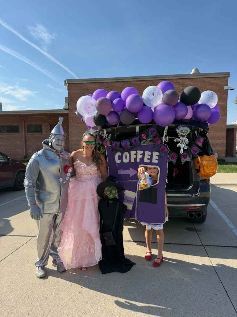 A group of four people dressed in costumes stands in front of a decorated car with balloons. The costumes include a silver tin man, a princess in a pink dress, a figure in a black cloak, and a young girl holding candy. The car is adorned with a sign reading 'Happy Coffee' and has Halloween-themed decorations.