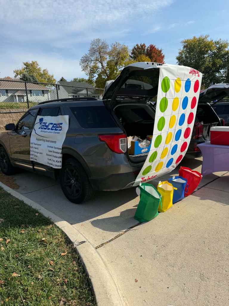 A grey SUV with its trunk open, displaying a Twister game backdrop and colorful containers in red, yellow, green, and purple on the ground. The vehicle has a sign advertising support for local events.