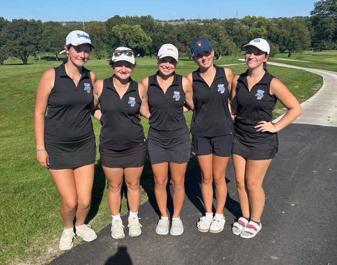 Five female golfers in matching black polo shirts and shorts pose together on a sunny day at a golf course.