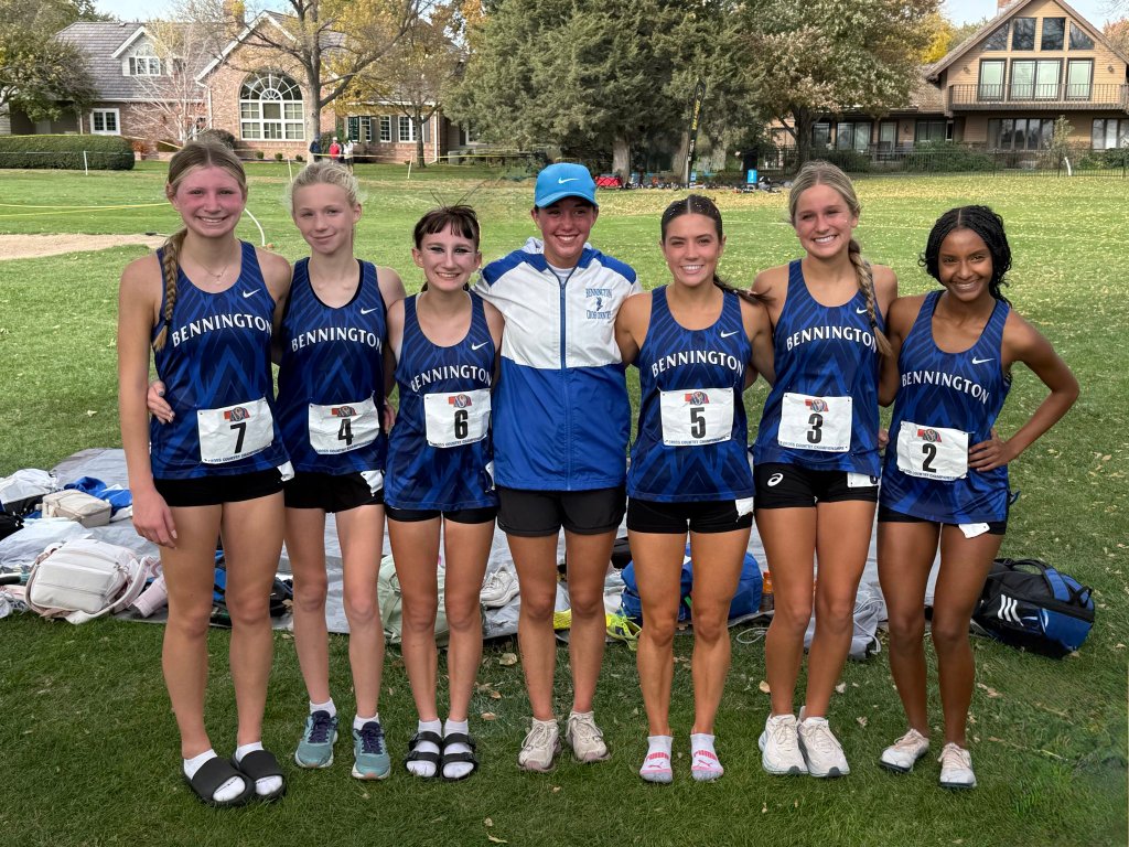 A group of six girls wearing blue running uniforms with 'Bennington' printed on them, posing for a photo with their coach in a grassy field. They are smiling, and there are backpacks and personal items in the background.