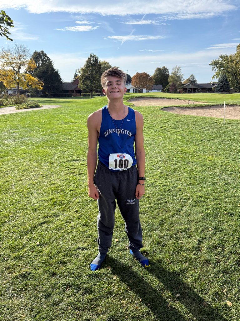 A young male athlete stands on a grassy field, wearing a blue sports tank top with 'BENNINGTON' printed on it and gray sweatpants. He has a race number '100' pinned to his outfit and is smiling at the camera, with a clear sky and trees in the background.