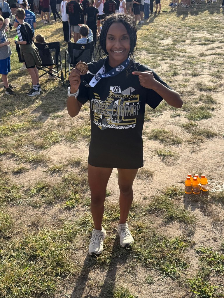 A young girl with braided hair smiles while holding a medal at a cross-country championship event. She wears a t-shirt celebrating the Nebraska state cross-country championships and is standing outdoors in a grassy area with spectators in the background.