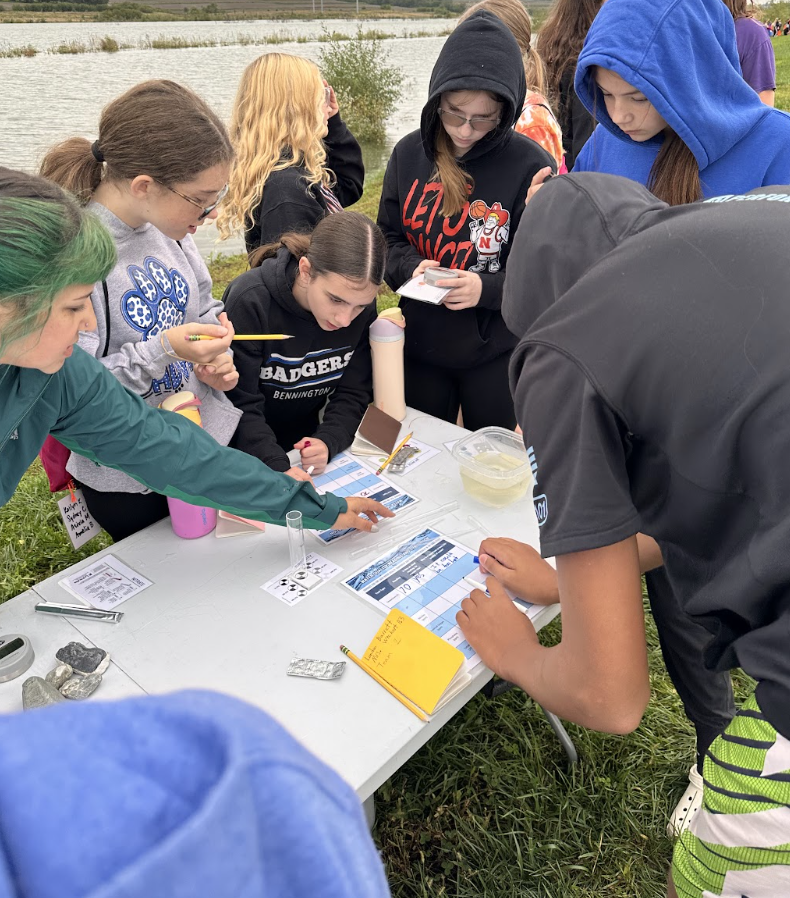 Group of students gathered around a table by a body of water, engaged in a science activity involving charts and testing materials.