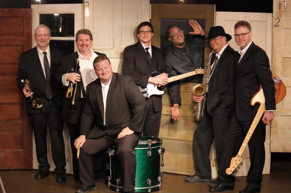 A band featuring seven members, dressed in black suits, posing together on stage. Instruments include a trumpet, saxophone, a drum set, and electric guitars. One member is playfully leaning out from a doorway.