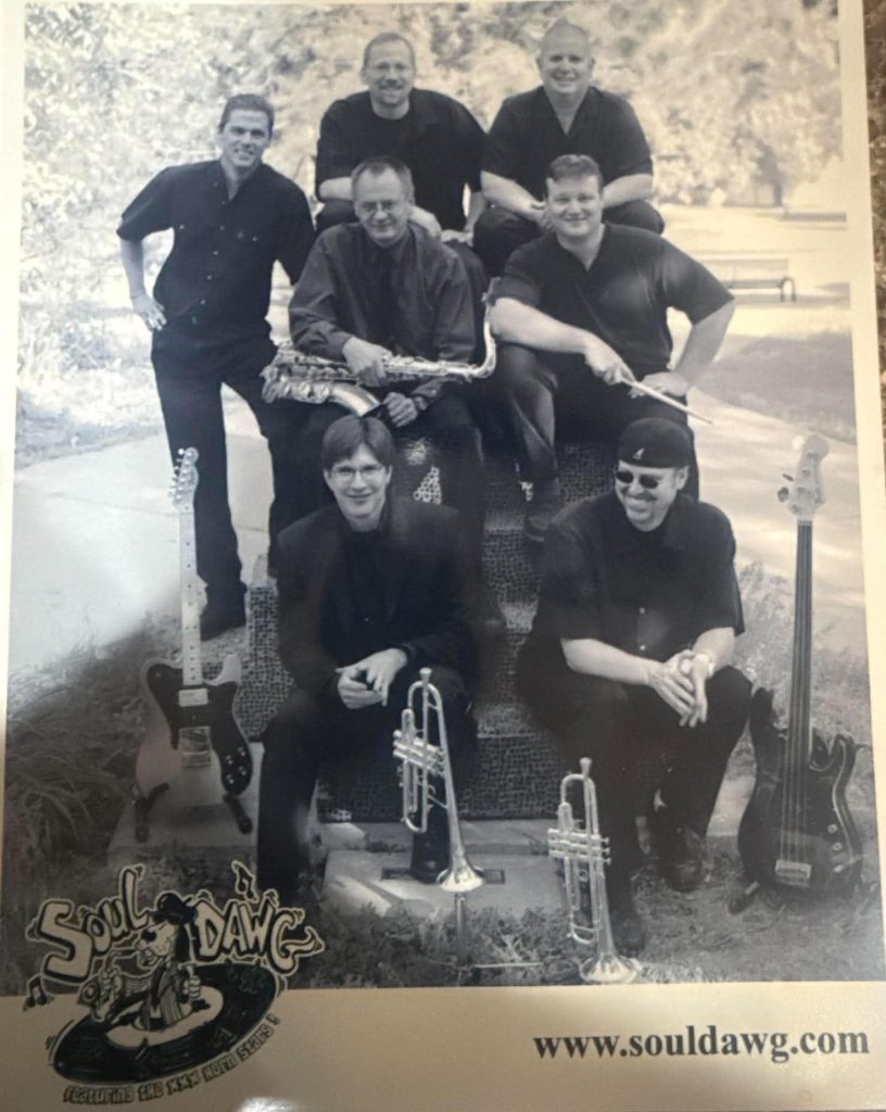 A black and white photo of a seven-member band, Soul Dawg, posing outdoors. They are arranged in two rows, with musical instruments including a guitar, saxophone, and trumpets visible. The band members are dressed in black attire.