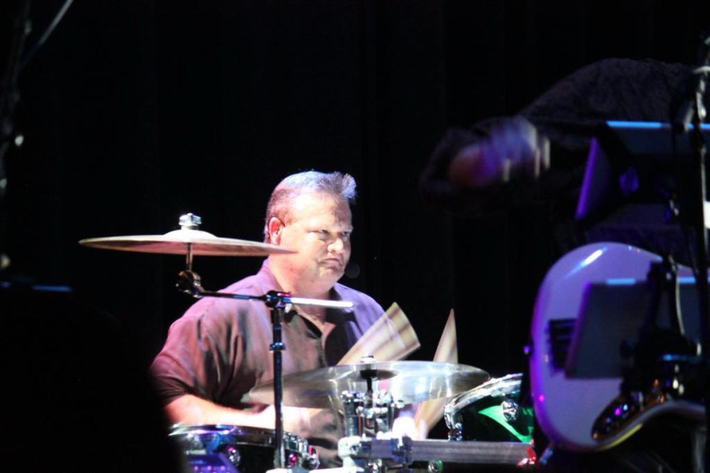 Drummer performing on stage, focused on playing a drum set with vibrant lighting in the background.