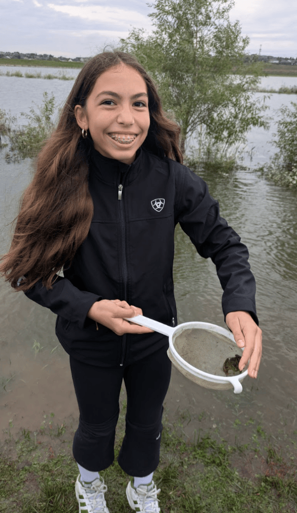 A girl with long brown hair smiles while standing by a body of water, holding a net with a small creature inside. She is wearing a black jacket and athletic pants.