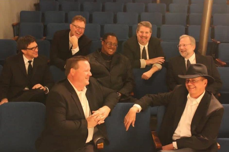 A group of eight men in formal black suits seated on theater chairs, smiling and posing for the photo.