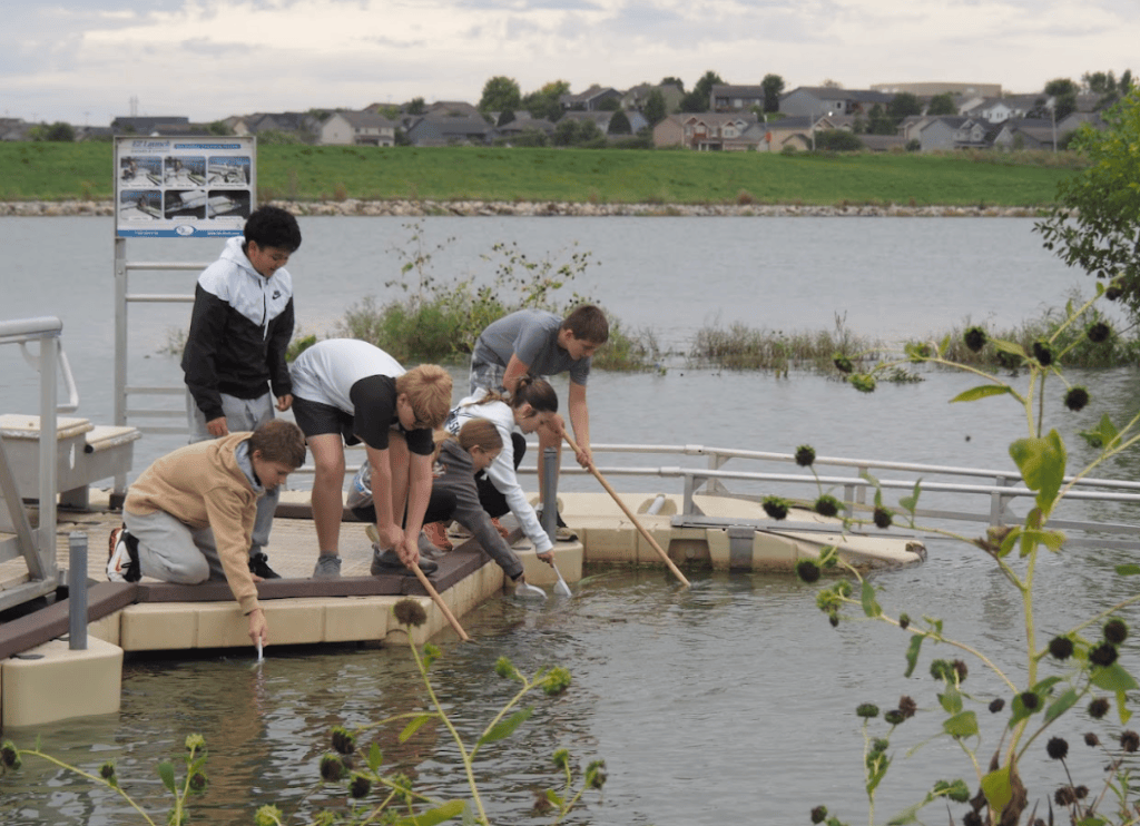 Group of children leaning over a dock, using sticks to explore the water of a lake, with houses and greenery in the background.