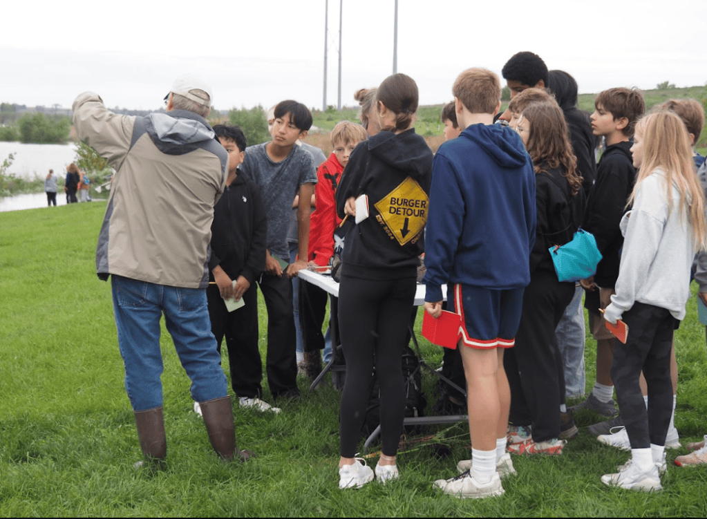 A group of children and an adult educator engage in an outdoor educational activity near a body of water, with the adult explaining something while the children listen attentively.