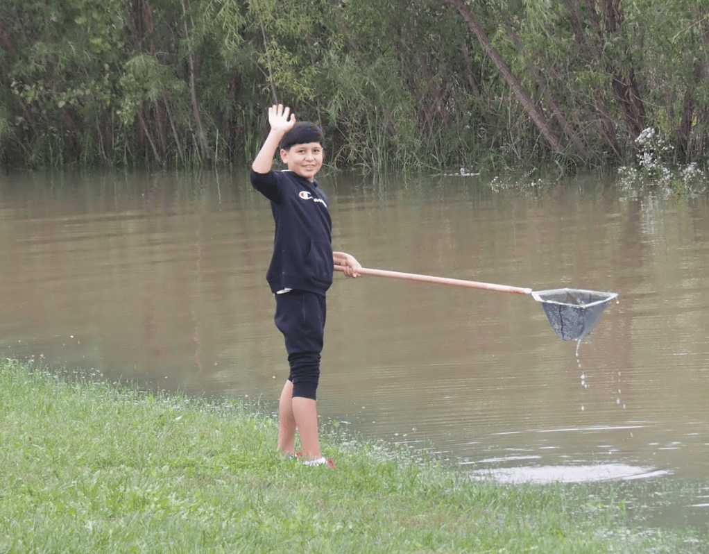 A young boy waves while standing by a pond, holding a fishing net with water splashing around.