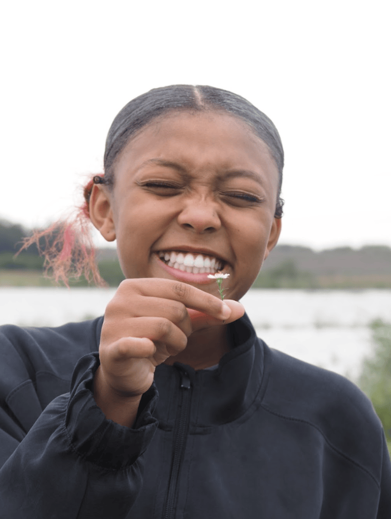A young person in a black jacket smiles broadly while holding a small flower, with a blurred natural background.