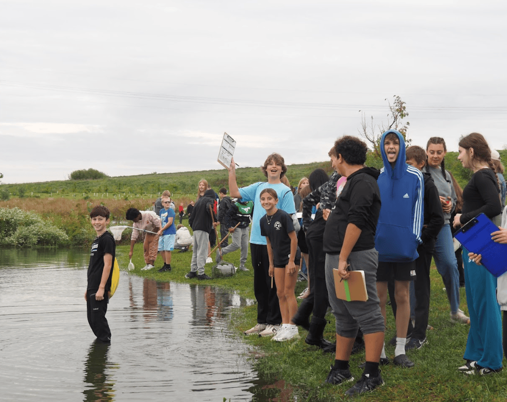 Group of students participating in an outdoor activity near a water body, some wading in the water and others smiling and engaging with each other.