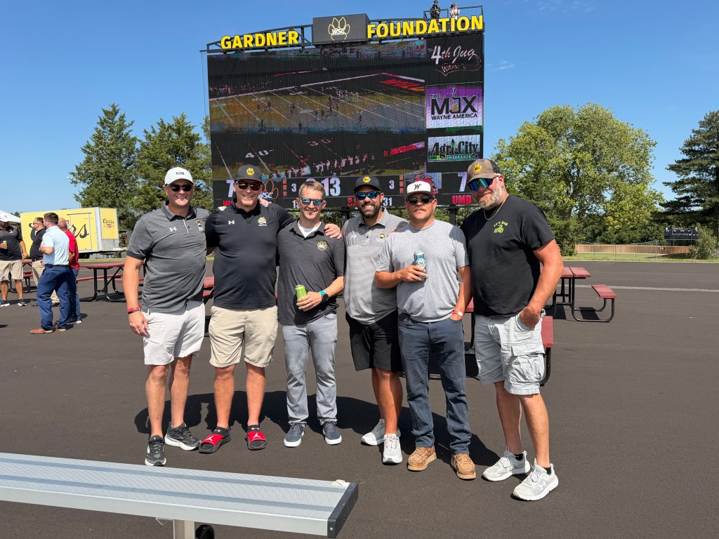 A group of six men posing for a photo outdoors, with a large digital screen displaying information in the background. They are wearing casual attire, including hats and sunglasses, and appear to be enjoying a sunny day at an event.