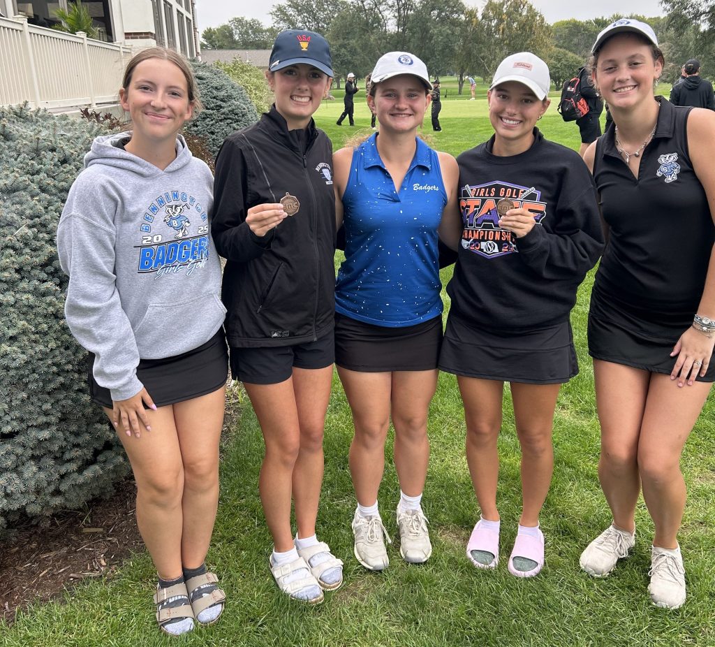A group of five female golfers standing together outdoors, smiling and posing for a photo. One player is holding a medal, and they are dressed in golf attire including skirts and caps.