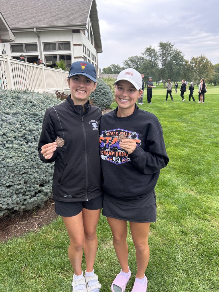 Two female athletes holding medals, standing on a grassy area with a clubhouse in the background.