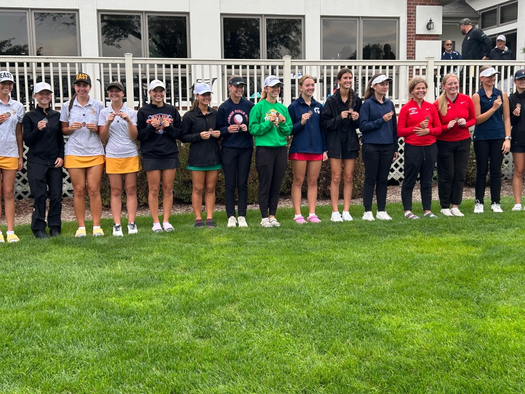 A group of female golfers standing in a row on green grass, proudly displaying medals or awards. They are dressed in various golf attire and hats, with a building in the background.
