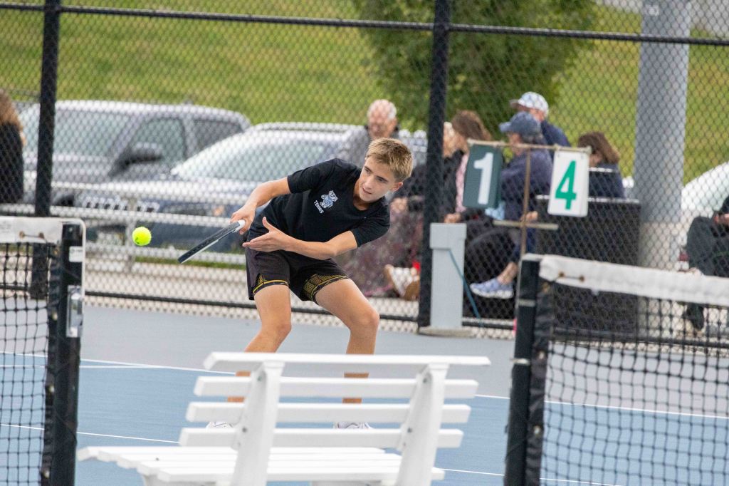 A young male tennis player preparing to hit a yellow tennis ball on a blue court, with spectators and cars visible in the background.