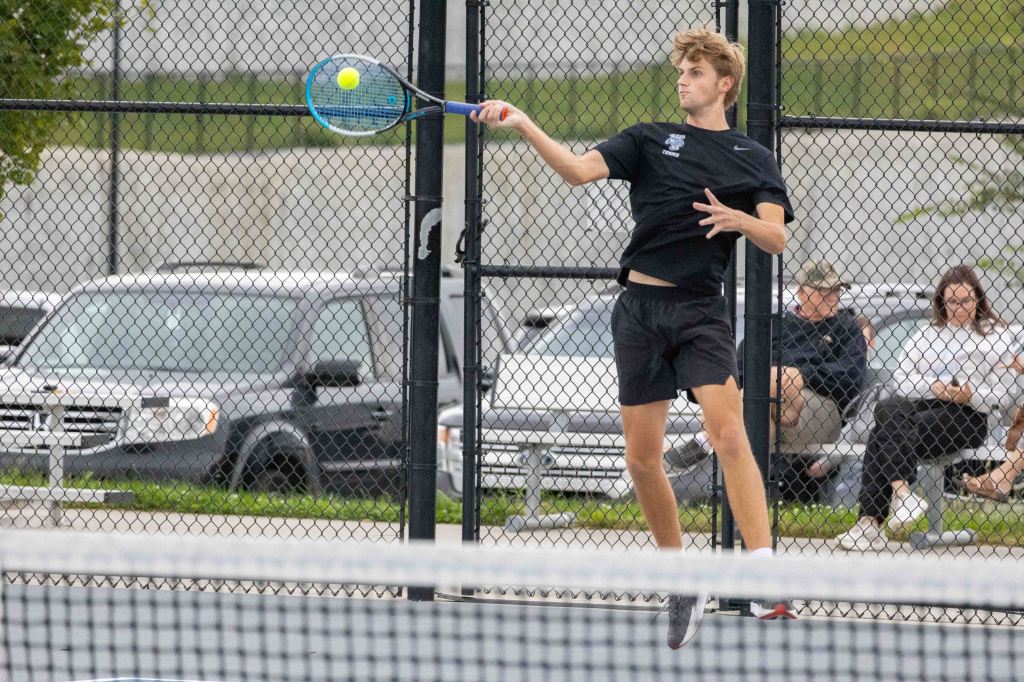 A young male tennis player is in mid-swing, preparing to hit a yellow tennis ball with a blue racket. The scene includes a tennis court surrounded by a chain-link fence, with spectators sitting in the background.