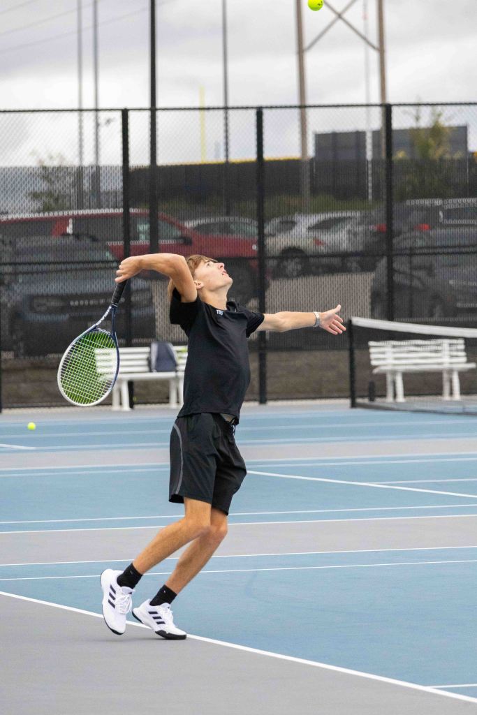 A young male tennis player serving a ball on a court, wearing a black shirt and shorts.