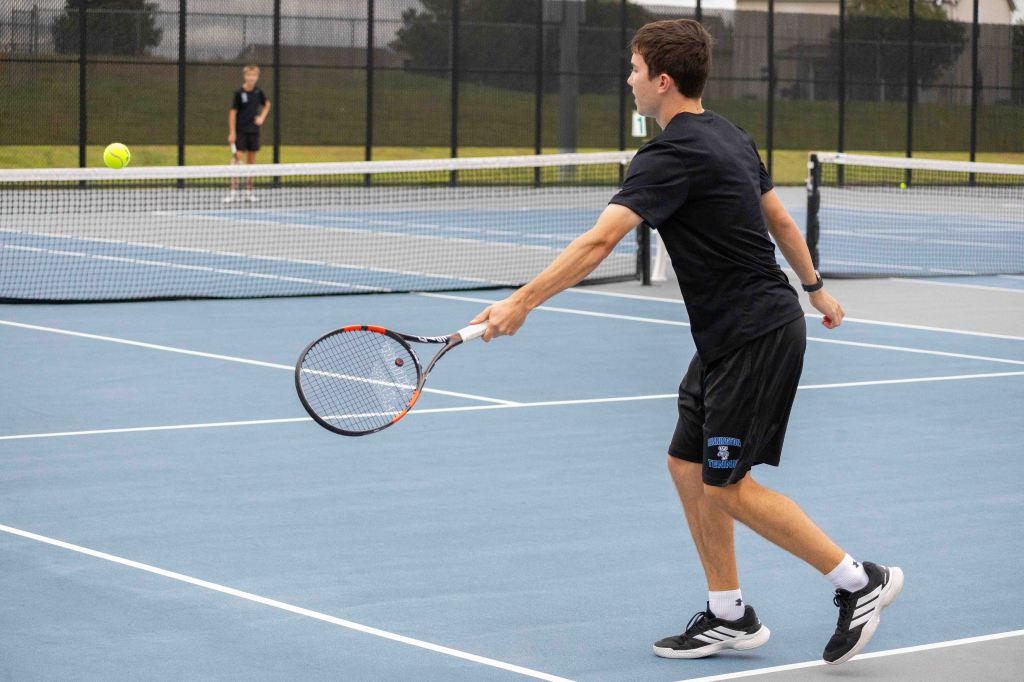 A young male tennis player hitting a forehand shot on a blue tennis court, with another player visible in the background.