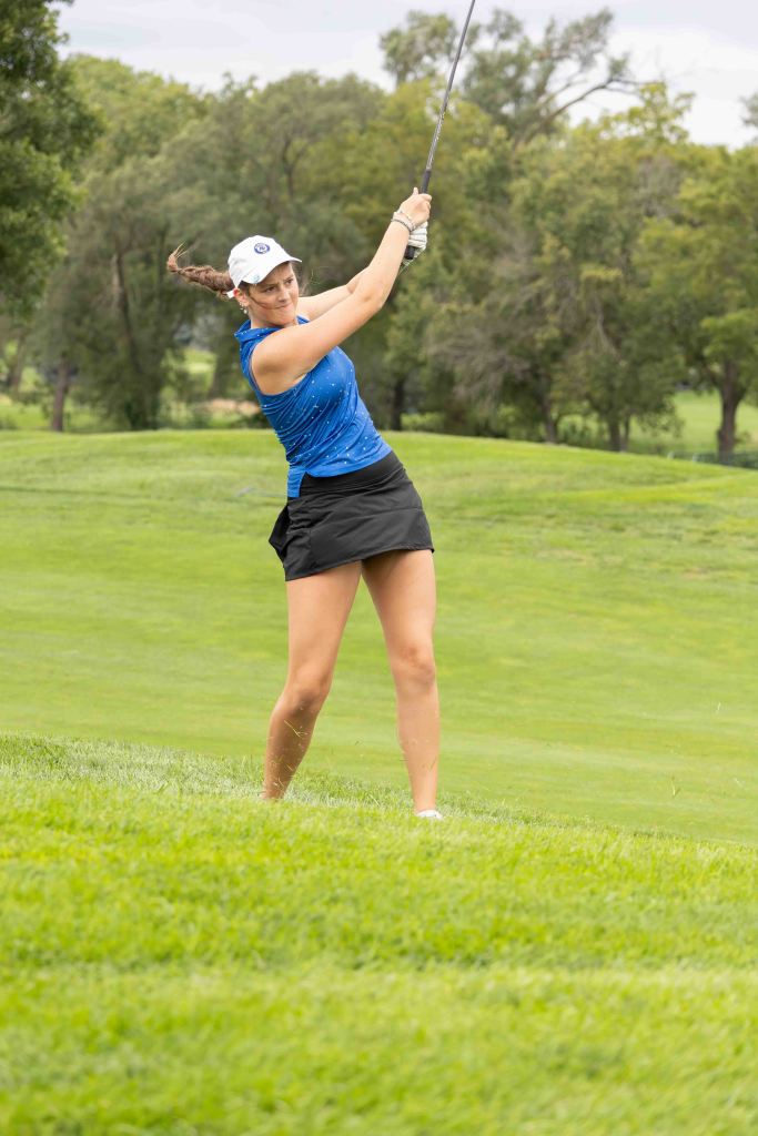 A female golfer in a blue shirt and black skirt is taking a swing with a golf club on a grassy course.