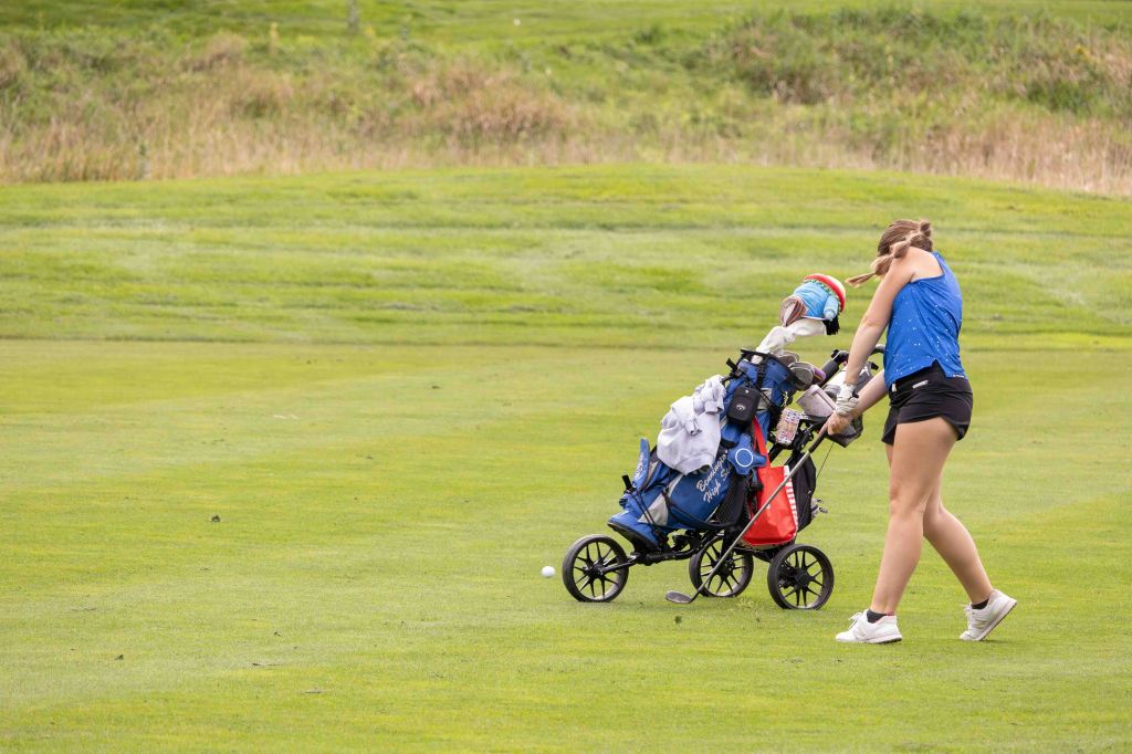 A woman pulling a golf pushcart with clubs and gear on a grassy golf course.
