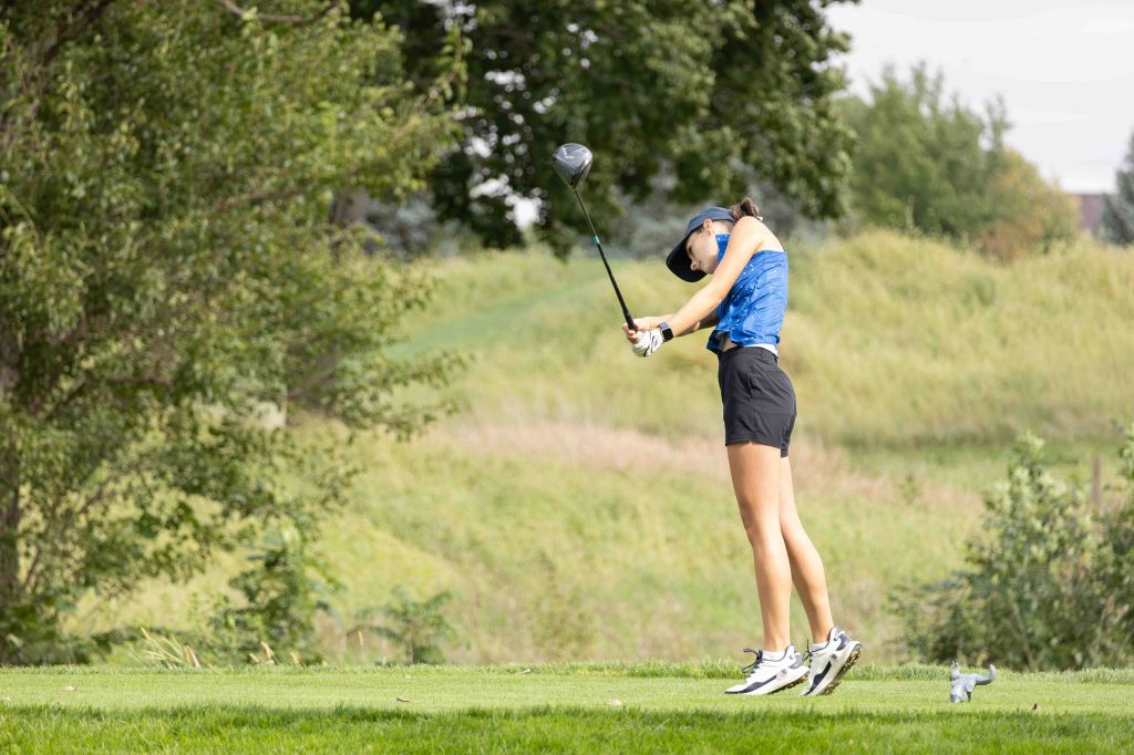 A golfer in a blue shirt and black shorts swinging a driver on a grassy course, surrounded by trees and a natural landscape.
